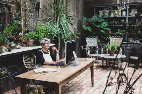 Preview: Senior woman wearing glasses, black top and white apron sitting at a wooden table, working on
