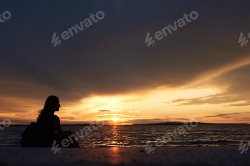 Preview: Silhouette of woman alone at water edge, enjoying beautiful seascape at sunset.