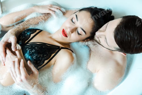 Preview: overhead view of handsome man and attractive brunette woman lying in bathtub with bath foam