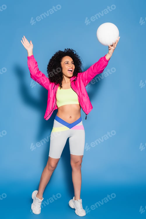 Preview: full length of amazed african american woman in pink jacket and crop top playing volleyball on blue