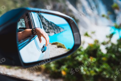 Preview: Female hand mirrored in the car side view mirror. Blue mediterranean sea and white rocks in