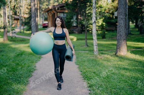 Preview: sportswoman wears cropped top and leggings, carries fitness ball and rolled up karemat