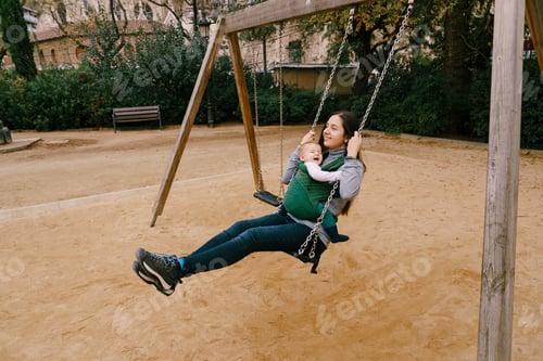 Preview: Smiling mom with little laughing baby in kangaroo backpack swinging on a swing in the park