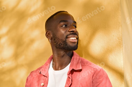 Preview: Smiling cool gen z Black hipster guy looking away standing at yellow wall.