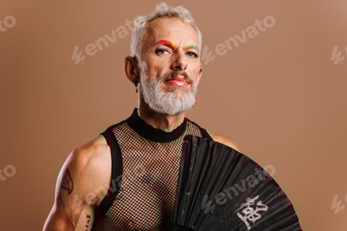 Preview: Bearded mature gay man with rainbow colored eyebrows holding hand fan against brown background