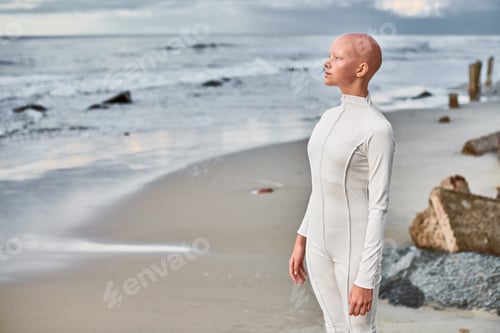 Preview: Full length portrait of hairless girl with alopecia in futuristic suit standing on seashore