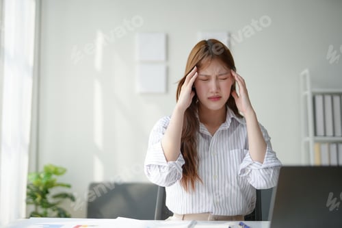 Preview: Woman experiencing a headache while working on her laptop in a modern office environment