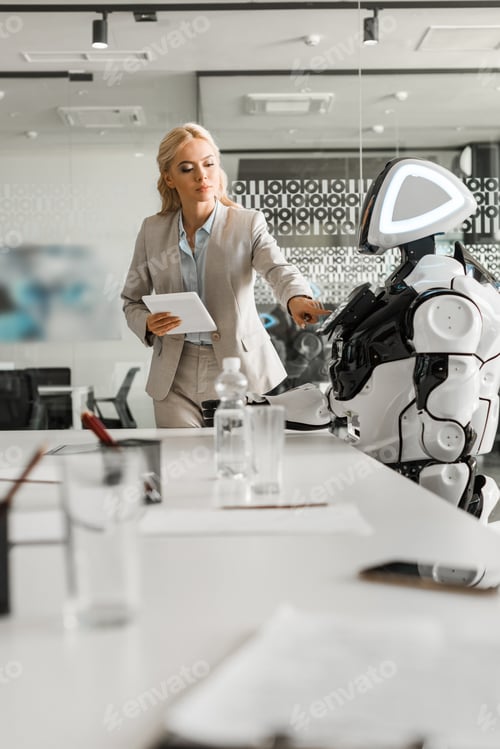 Preview: selective focus of attractive businesswoman operating robot while holding digital tablet