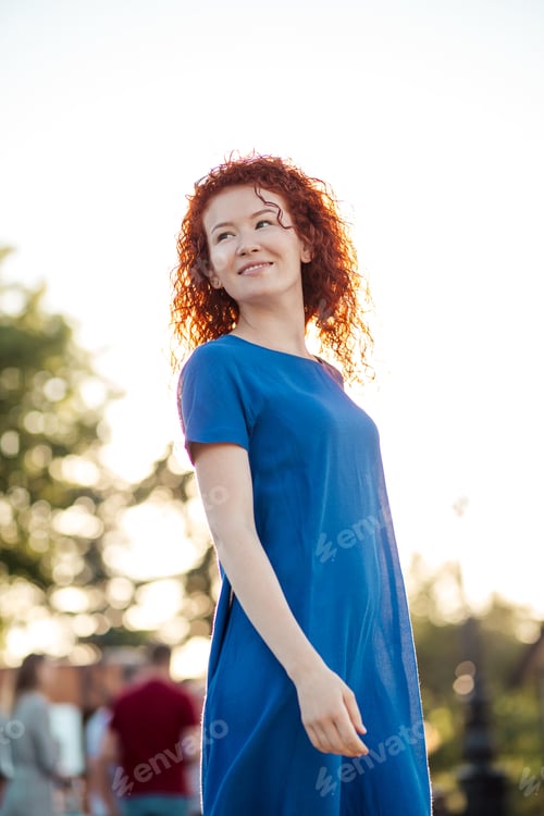 Preview: Young woman with waved curly red hair, in blue dress, in park.