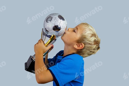 Preview: Young soccer player in blue jersey holds winners cup after the goal, isolated at light background