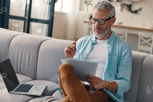 Preview: Thoughtful senior man checking the papers with serious face while sitting on the sofa at home