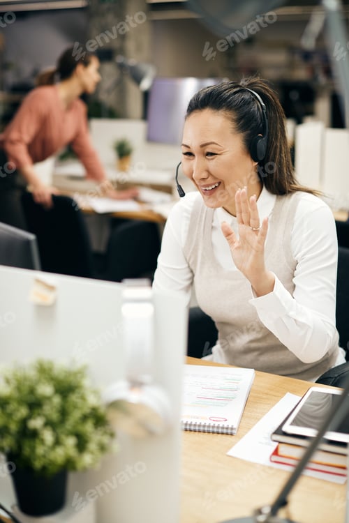 Preview: Happy Asian businesswoman greeting someone while having video call in the office.