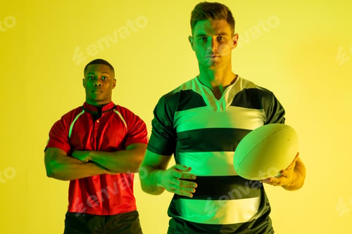 Preview: Portrait of diverse male rugby players with rugby ball over yellow lighting