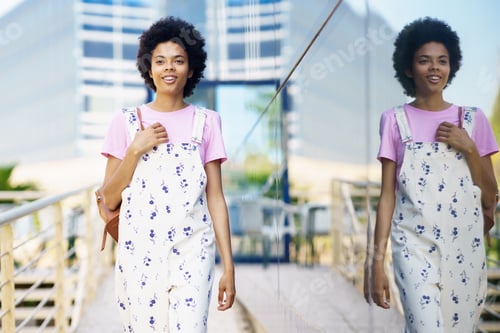 Preview: Cheerful African American woman in casual clothes walking between modern buildings smiling.