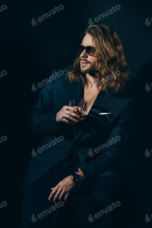 Preview: Handsome bearded man in fashionable suit holding glass of alcoholic beverage and looking away on