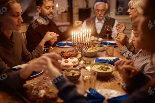 Preview: Close up of extended family holding hands and praying while celebrating Hanukkah at dining table.