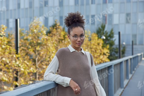 Preview: Horizontal shot of beautiful curly haired young woman wears round spectacles shirt and knitted vest