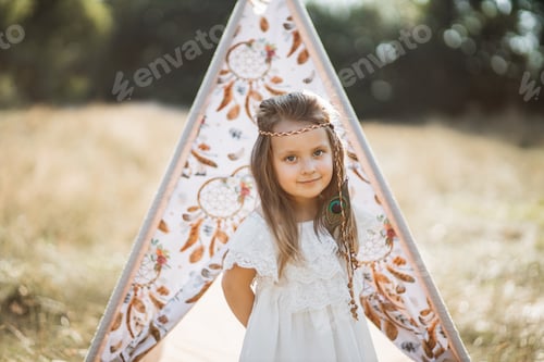 Preview: Pretty small girl, wearing stylish white dress and feather hair accessories standing near teepee