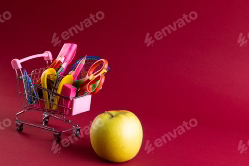 Visualização: Vista de perto do carrinho de compras em miniatura com materiais escolares, maçã e espaço de cópia em fundo vermelho
