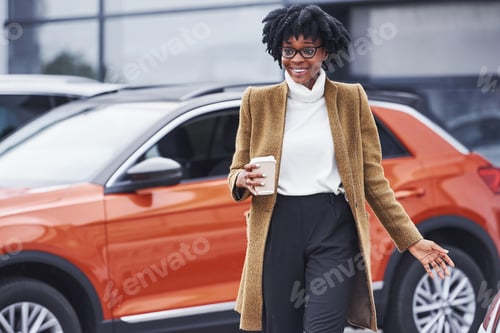 Preview: Young african american woman in glasses and with cup of drink stands outdoors near modern car