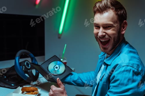 Preview: selective focus of handsome and smiling man holding compact disk with video game and showing like