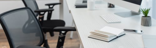Preview: notebook, mobile phone and flowerpot near keyboard on desk near blurred office chairs, banner