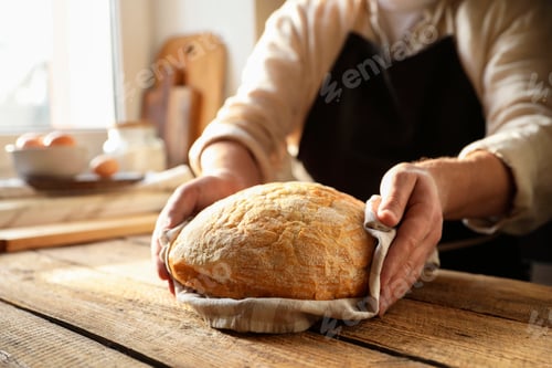 Preview: Man holding loaf of fresh bread at wooden table indoors, closeup