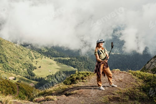 Preview: Young woman in cap taking selfie on smartphone on hike against backdrop of mountain valley and cloud
