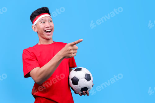 Vista previa: Energetic Male In Red Jersey And Headband Pointing To The Side With Soccer Ball In Hand