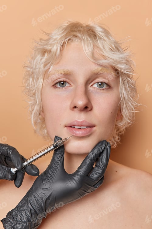 Preview: Serious blonde woman gets cosmetic injection in lips at beauty salon stands with bare shoulders