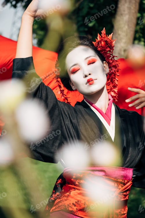 Preview: selective focus of tree branches and beautiful dancing geisha with red cloth on background in