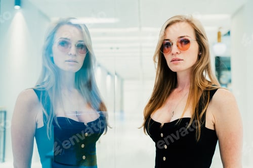 Preview: Gorgeous red haired young woman in sunglasses stands indoors reflected in glass. European model