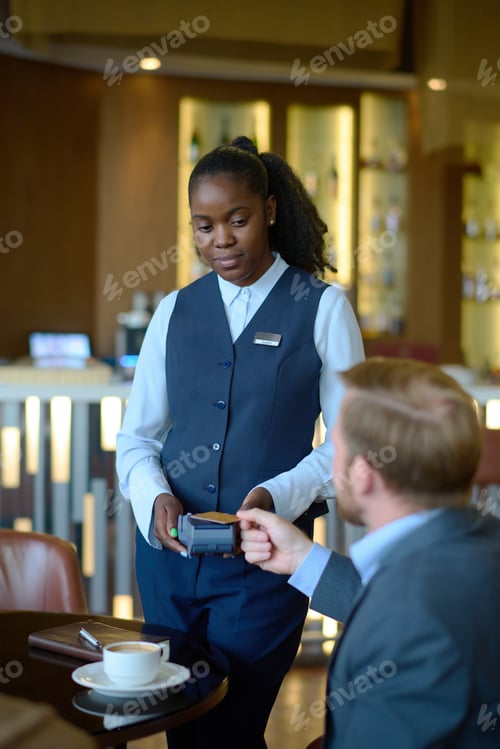Preview: Young worker of hotel with payment terminal standing in front of businessman