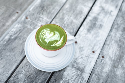 Preview: Cup of fresh green matcha latte beverage with latte art on foam. Background of wooden table