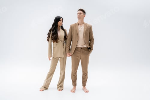 Preview: full length of barefoot multiethnic couple in trendy suits holding hands while standing on grey