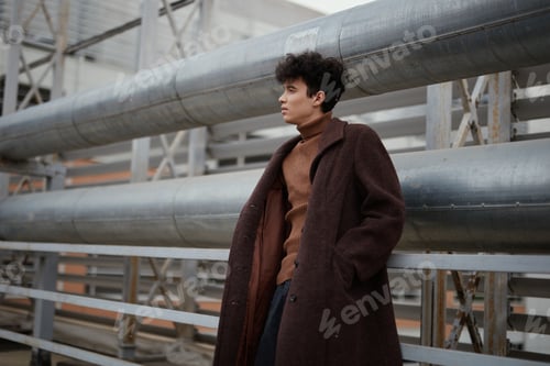 Preview: Fashionable man in a brown coat and sweater leaning against a metal railing in front of a large pipe