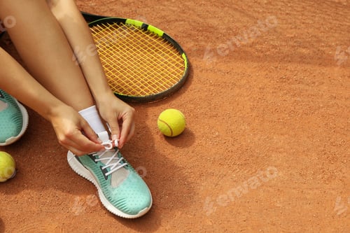 Preview: Woman tying shoelaces on clay court with racket and tennis balls