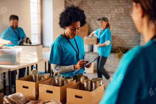 Preview: Black female volunteer making list of food supplies while working at humanitarian aid center.