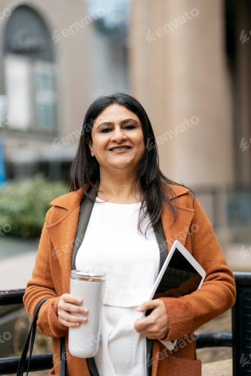 Preview: Smiling Indian business woman holding digital tablet, bottle of water looking at camera walking on t