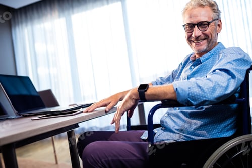 Preview: Sitting senior man typing on laptop at modern office desk, with wheelchair, copy space