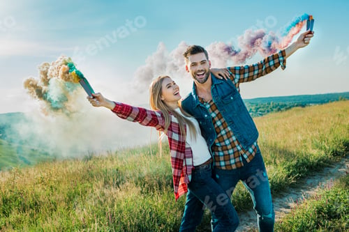 Preview: young couple holding colorful smoke bombs on rural meadow