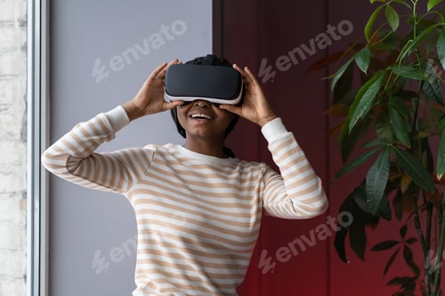 Preview: Amazed african american woman testing modern high-tech VR helmet, getting 3D experience at home