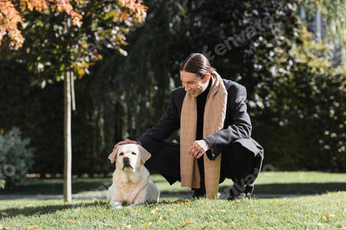 Preview: full length of tattooed man in stylish coat and scarf smiling while cuddling labrador in park