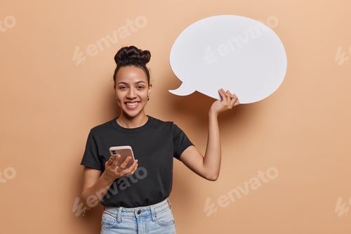 Preview: Horizontal shot of positive cheerful Latin woman with hair bun holds mobile phone for online