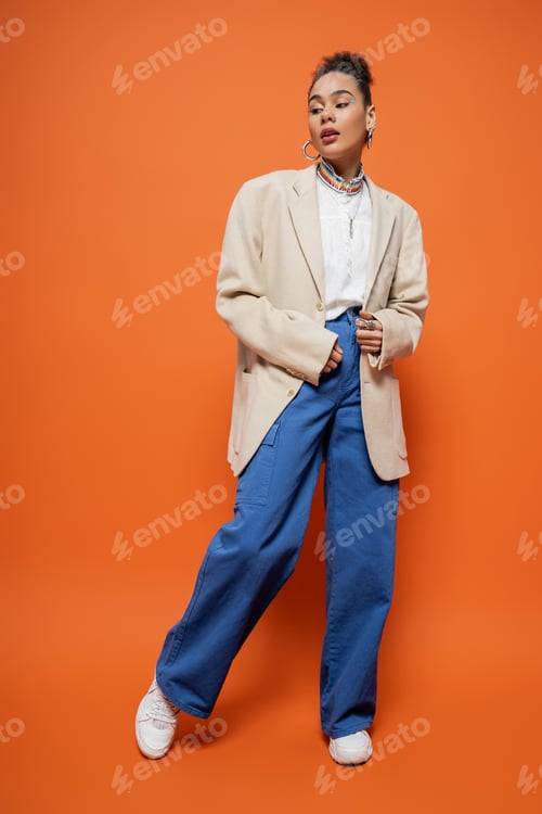 Preview: pretty african american model in stylish beige blazer and blue pants posing on orange background