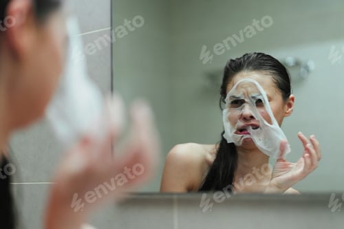 Preview: Young Asian woman applying facial mask in bathroom, looking in mirror with a surprised expression