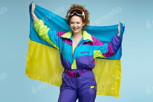 Preview: Smiling redhead woman holding ukrainian flag wearing colorful retro ski suit and goggles