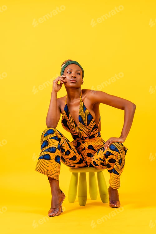 Preview: Young African woman in traditional dress on yellow background, sitting, commercial photography
