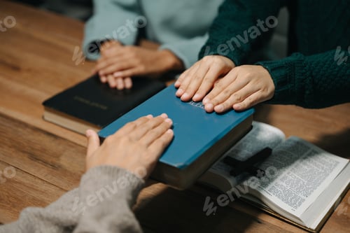 Preview: Group of Christians sit together and pray around a wooden table with blurred open Bible pages