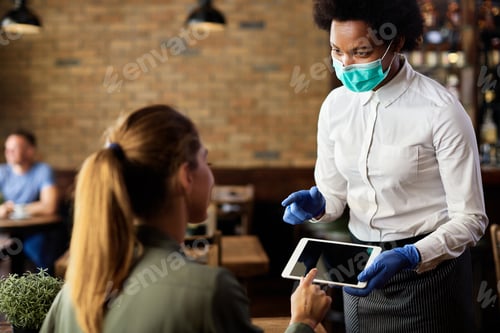 Preview: Black waitress wearing protective face mask while taking order on touchpad from her customer.
